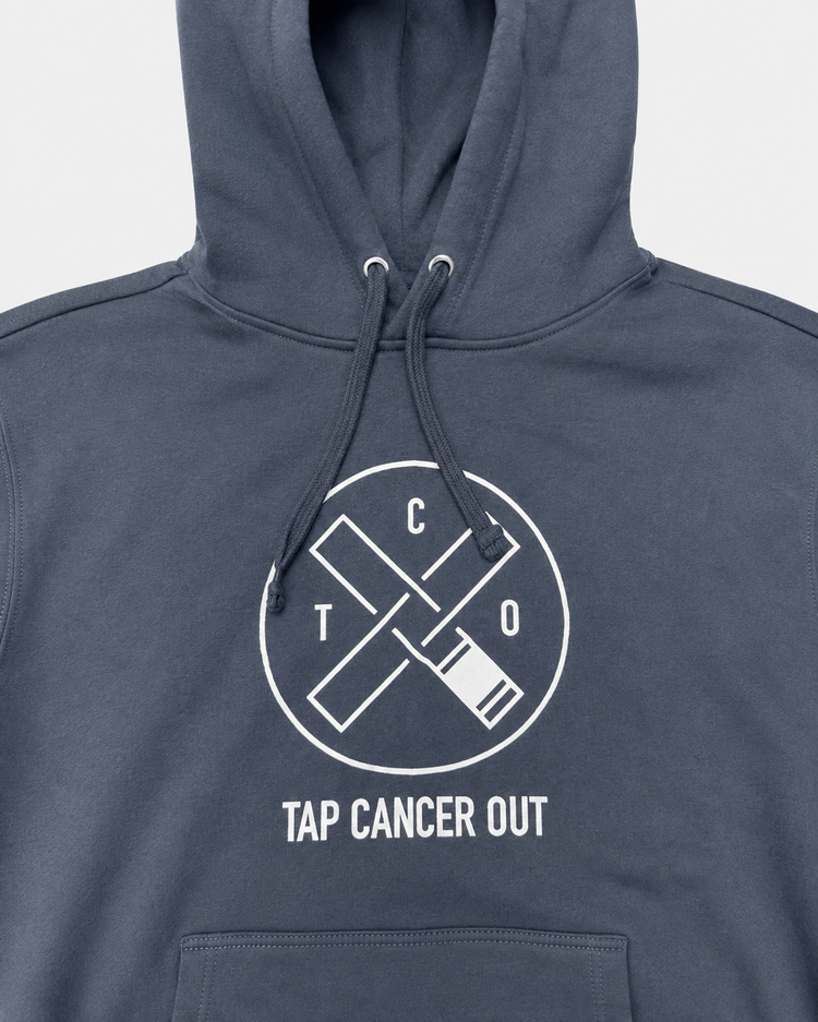 A close-up of a storm blue hoodie featuring a white circular graphic with crossed belts and the text 'TAP CANCER OUT' on the chest.